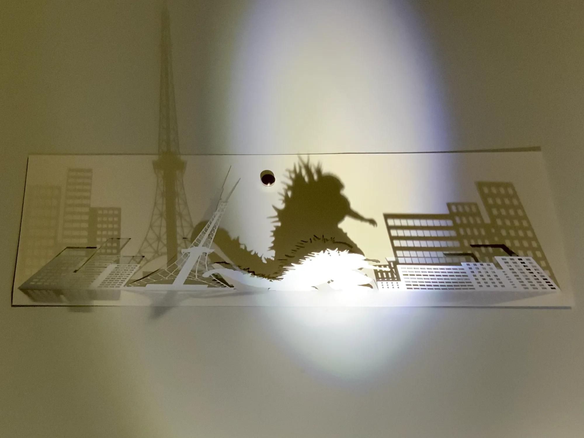 光と影で街並みを描く影絵作品。塔と高層ビルのシルエットを紙の切り絵で構成したペーパークラフト